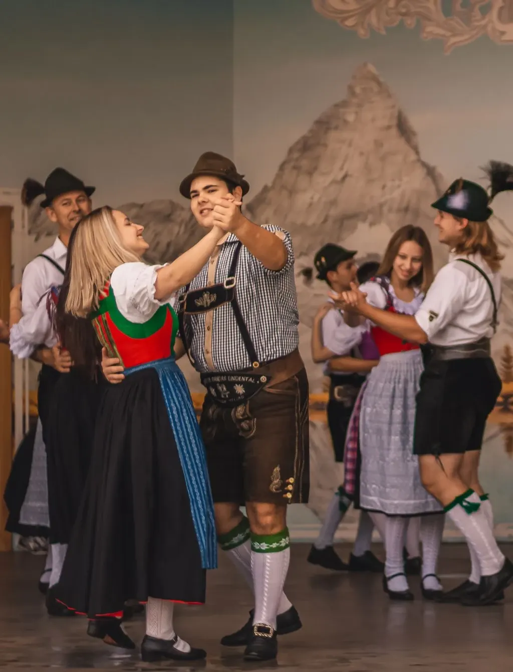 Casais dançando em trajes típicos tiroleses, com os Alpes pintados ao fundo